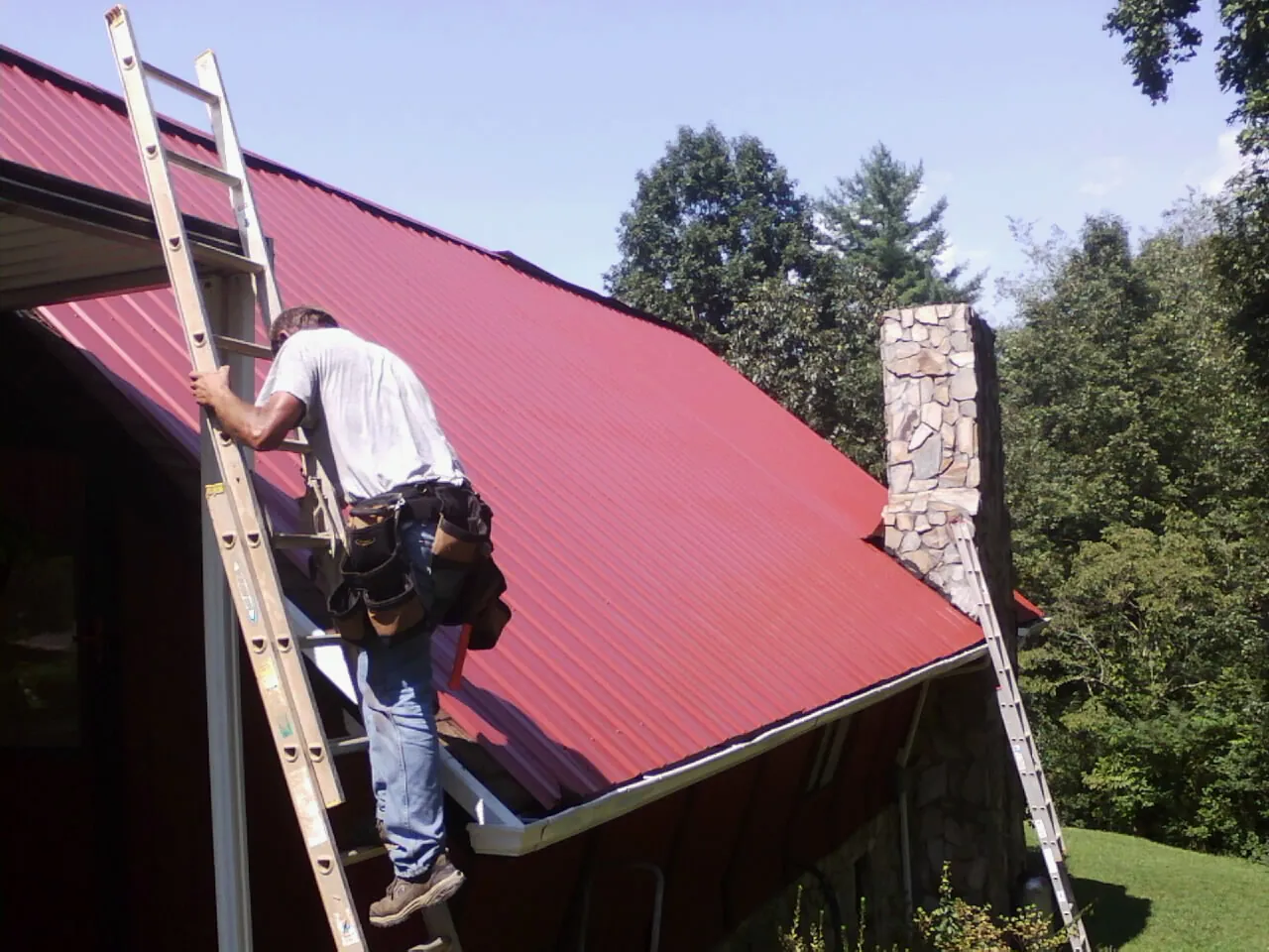 Roofer on a ladder beside a red metal roof and stone chimney — Soffit & Fascia Repair by Roofing Brothers in Weber City